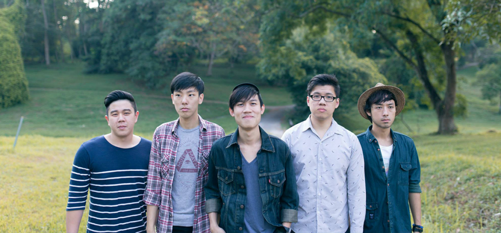 Five young men stand together in a grassy park, posing for the camera with trees in the background