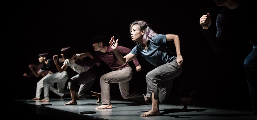 Group of dancers on a dim stage mid-mountainous sprint-like pose, bending forward with arms extended, focused expressions under dark lighting.
