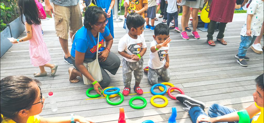 Adults and children kneel and stand around a ring-toss game on a wooden deck, colorful rings scattered on the ground.