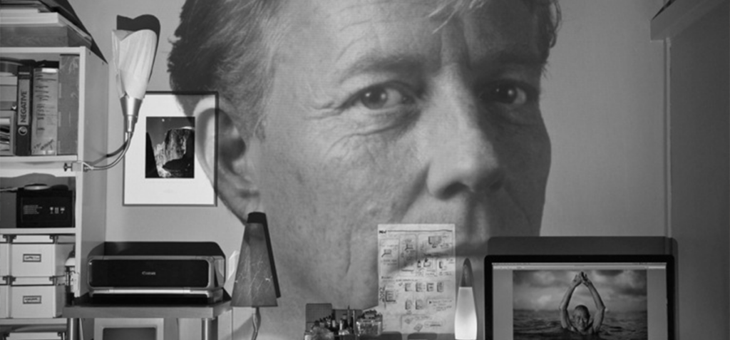 Large grayscale wall mural of a man's face dominating a cluttered home office with a printer, shelves, and computer monitor.