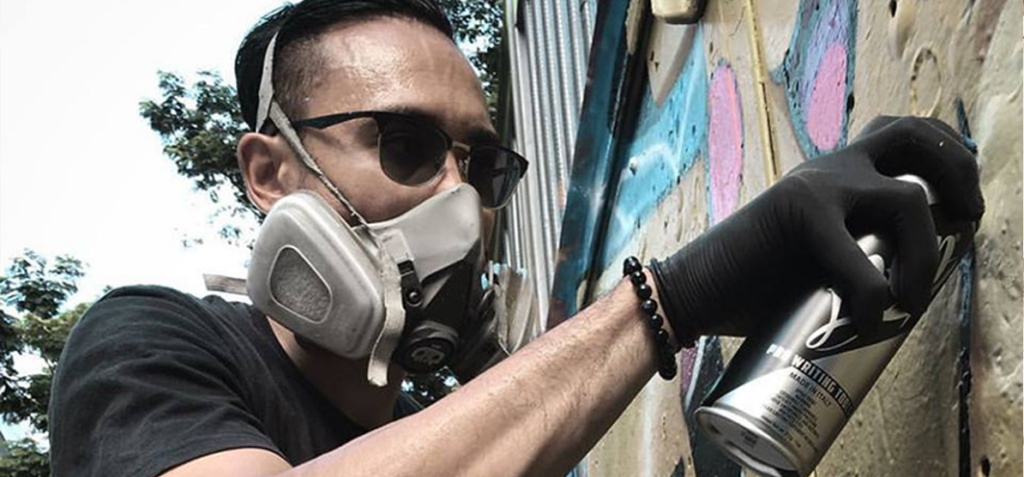 Person wearing a respirator mask and black gloves spray-painting graffiti on a wall.