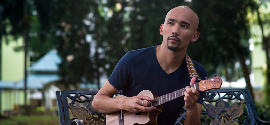 Man playing a small wooden ukulele on a decorative park bench outdoors, wearing a dark blue shirt and looking to the side.