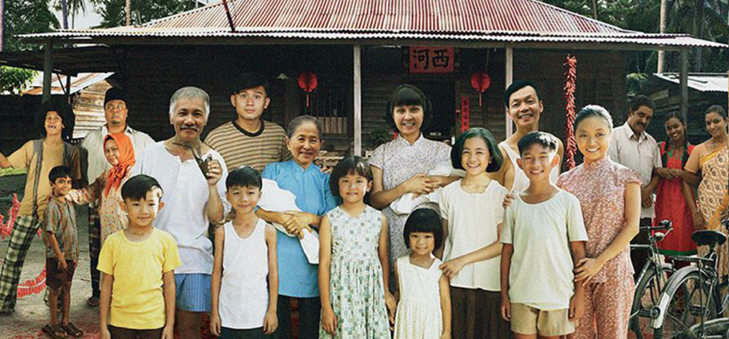 Multigenerational group photo in front of a wooden house with red lanterns; children, adults and seniors pose together, some holding babies.