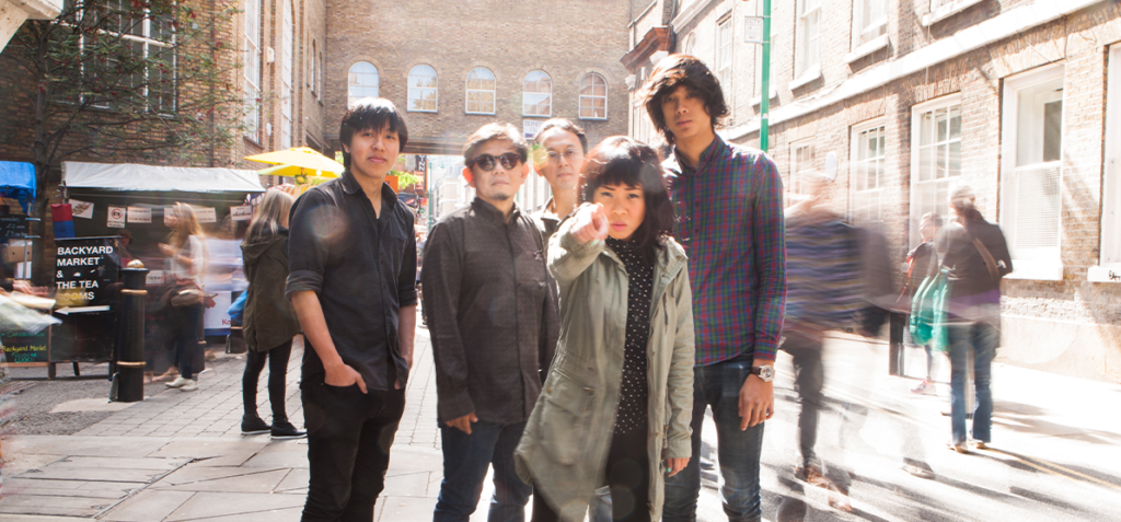Five young adults pose on a sunny city street with brick buildings; blurred pedestrians pass by behind them.