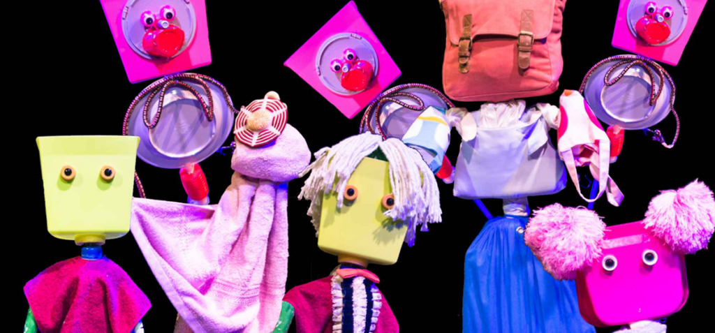 Whimsical puppets with square heads and bright outfits on a black stage, many bags and props around them.